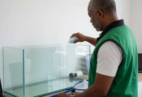 Pet store employee adding water conditioner to a large aquarium with fishing net and gravel on the counter