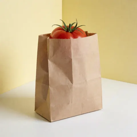 A fresh red tomato with a green stem sits inside a brown biodegradable paper bag on a white surface with light yellow walls.