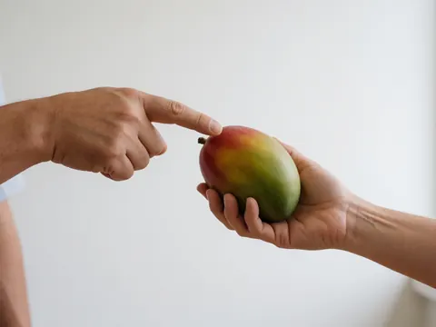 Adult hand pointing at a ripe mango with realistic skin against a clean white background, showing fresh fruit detail in a produce store.