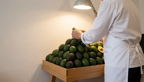 Seller arranging avocados in a pyramid on a wooden table with warm lighting and white wall, copy space