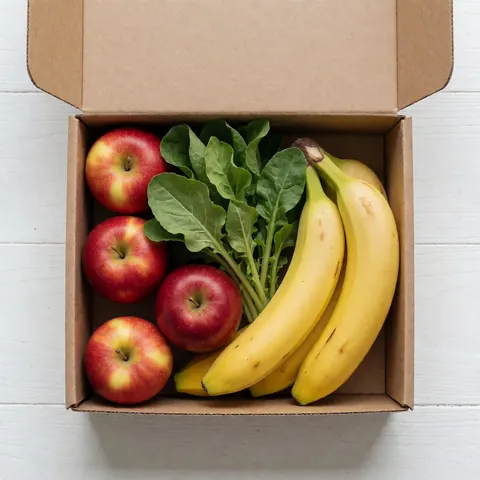 Cardboard box filled with fresh apples, bananas, and green vegetables arranged on a clean white table for a grocery store display
