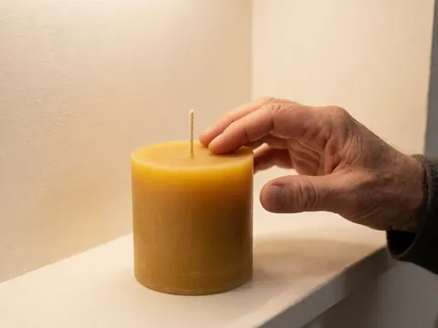 Adult hand touching a beeswax candle on a white shelf with warm soft walls, ideal for artisanal perfumery.