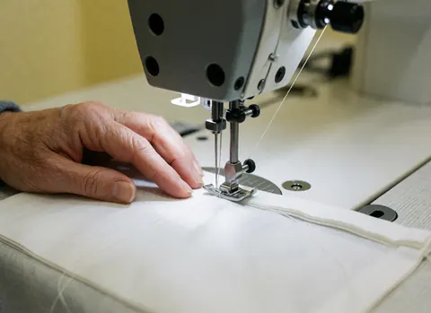 Close-up of an adult hand guiding white medical fabric under the needle of an industrial sewing machine with a pale yellow wall in the background.