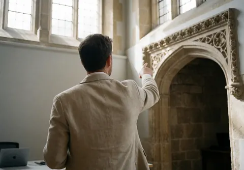 Tour guide pointing to a carved stone arch in a historic museum with natural light, gray walls, and tall windows
