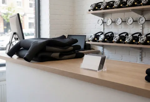 Dive counter displaying folded wetsuits and regulators on shelves with certification cards against a white brick wall and natural light
