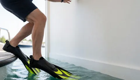 A leg wearing black and yellow diving fins emerging from the water near a boat, with a white wall in the background at a diving school.