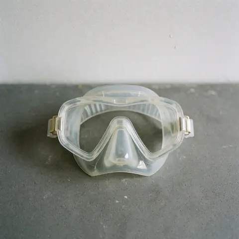 Diving mask with clear lens and silicone border on textured gray surface against white wall with natural lighting