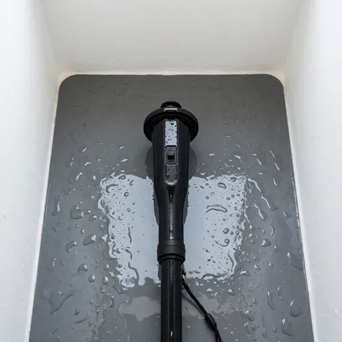 Black underwater torch resting on a wet gray rubber mat with white walls, suitable for diving equipment rental
