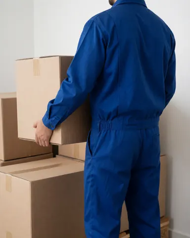 A logistics worker in a blue uniform lifting a cardboard box in a clean warehouse with white walls.