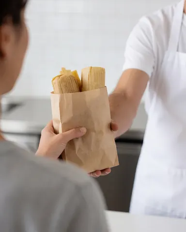 A customer holds a paper bag of tamales handed to them by a vendor in a butcher shop with white tiled walls.