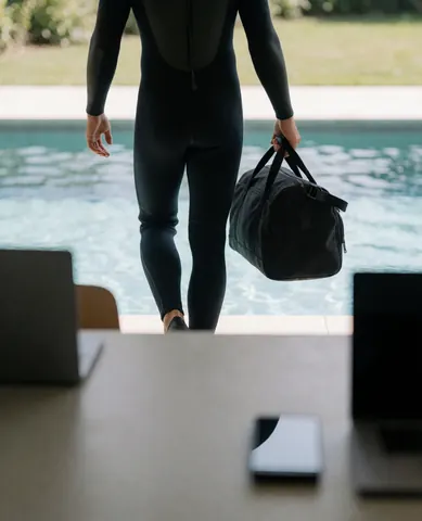 Diving instructor in black wetsuit carrying gear bag walking toward pool from behind in natural light