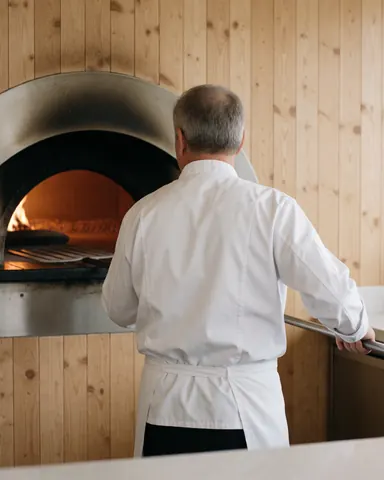 Chef standing with back to camera in front of a wood-fired oven with visible flames and wooden walls in a realistic pizzeria setting