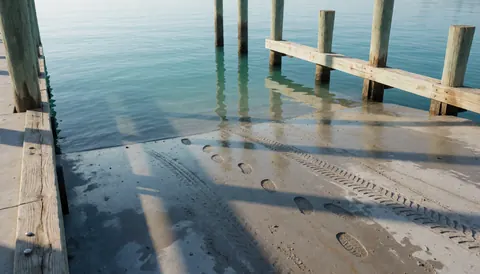 Wet concrete ramp with tire and foot tracks, wooden posts, and turquoise water, suitable for a dive school entrance.