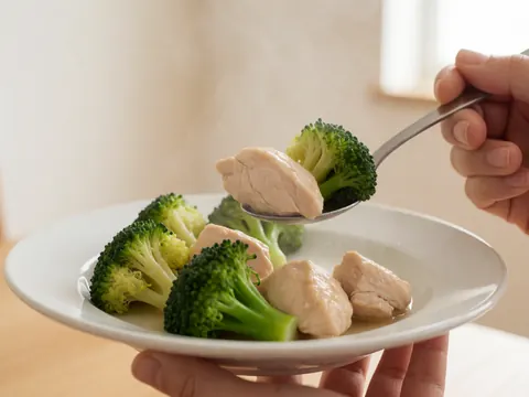 Close-up of a hand holding a spoon with chicken and broccoli over a white plate, representing hot meals for seniors.