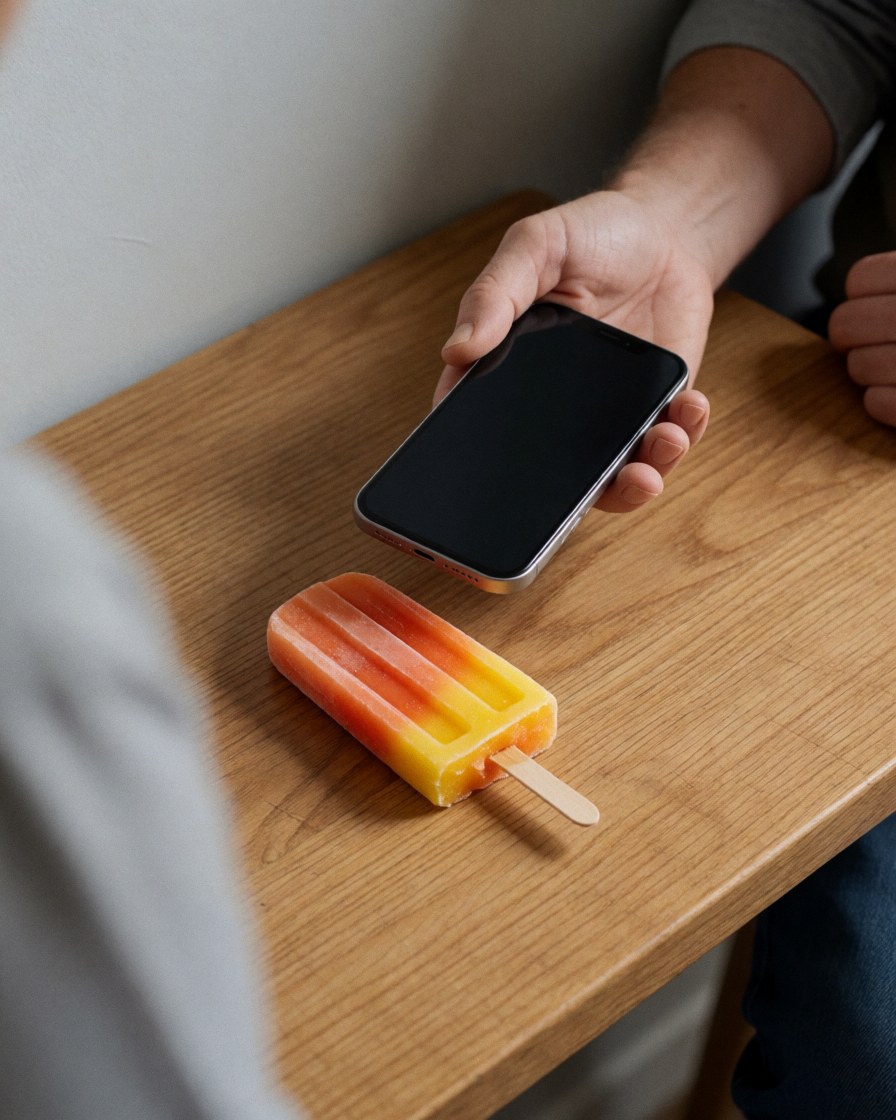 A hand holding a smartphone above a fruit popsicle on a wooden table in an ice cream shop with warm gray walls.