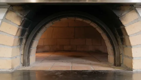 Interior view of a pizzeria featuring a large brick oven with arched openings and tiled flooring, providing a warm background for text.