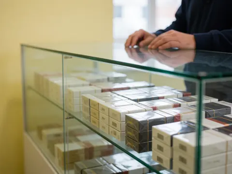 Adult hands arranging cigarette packs in a glass display case at a gas station with a pale yellow wall