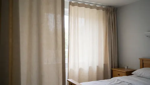 Senior care room with blackout curtains, white bed linens, wooden nightstand, natural light, and light gray walls